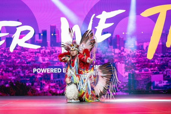 Native American dancer in traditional regalia performing on stage in front of a city skyline.